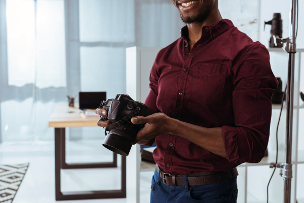 cropped shot of smiling african american photographer holding photo camera in hands in studio